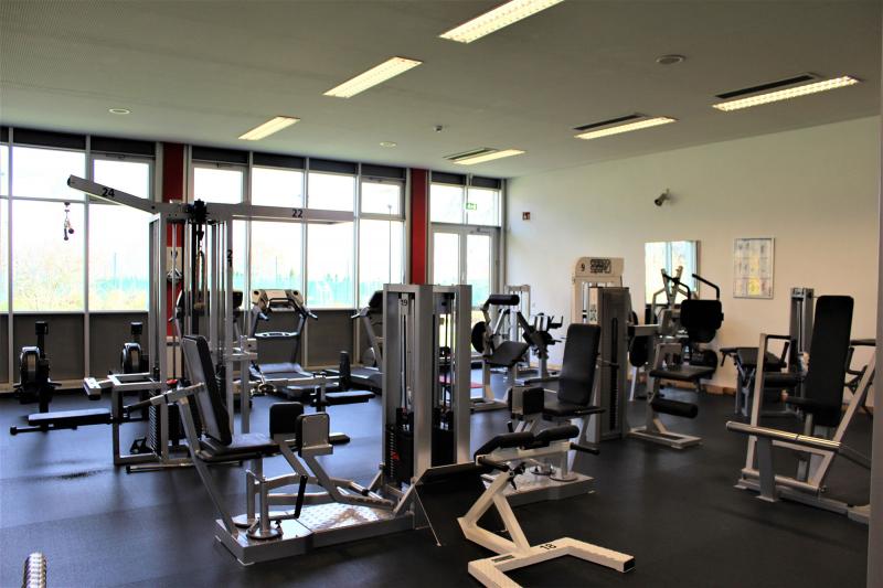 Fitnessstudio mit Geräten zum Muskelaufbau und im Hintergrund sind Laufbänder und Rudermaschinen.